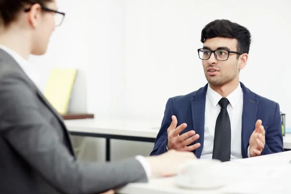 A young professional in a suit gestures while speaking in a bright office interview or meeting.