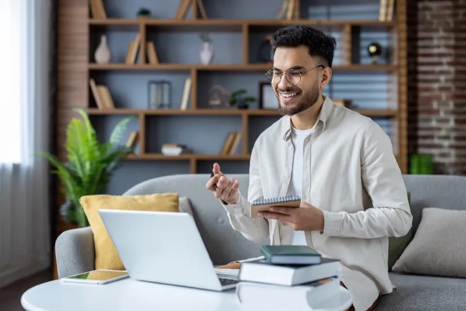 A young professional on a video call from a home office, taking notes with a laptop and books on the table.